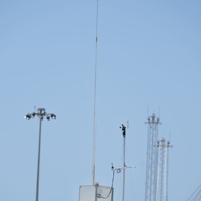 A rooftop with various antennas and communication equipment under a clear blue sky at the Port Hueneme Little League stadium. Multiple light poles and tall structures are visible in the background, capturing the essence of a sunny day at the ballpark.