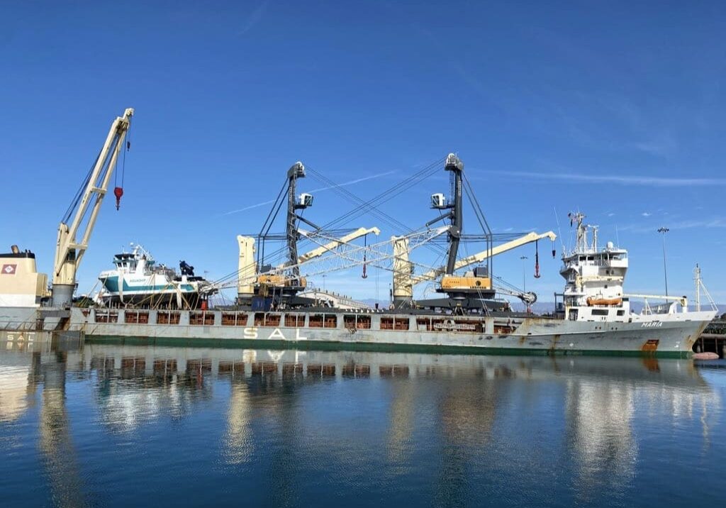 A large cargo ship docked at a harbor with cranes alongside, under a clear blue sky. Various machinery and equipment are visible on the deck in this bustling scene of Year End 2021, capturing the essence of Dock Talk.