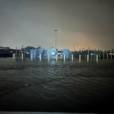 A dark, flooded outdoor area with water covering the ground and surrounding several tall, lit-up structures equipped with shoreside power.