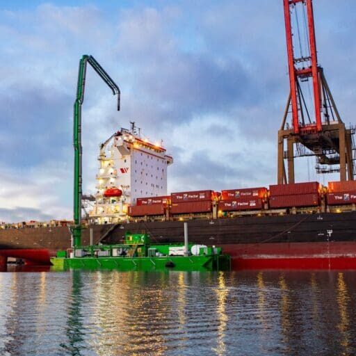 A cargo ship is docked at a port with cranes unloading shipping containers, benefiting from the $3.935 million state grant for Port of Hueneme as Governor ramps up California Port Data Partnership Program. A green barge is moored alongside the ship, under a cloudy sky with calm waters.