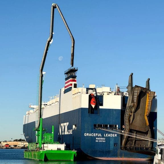 A large blue and white cargo ship is docked at the Port of Hueneme, sporting a crane on its deck. Under the clear blue sky, with the moon in view, this bustling port benefits from a $3.935 million state grant as part of Californias enhanced Port Data Partnership Program.