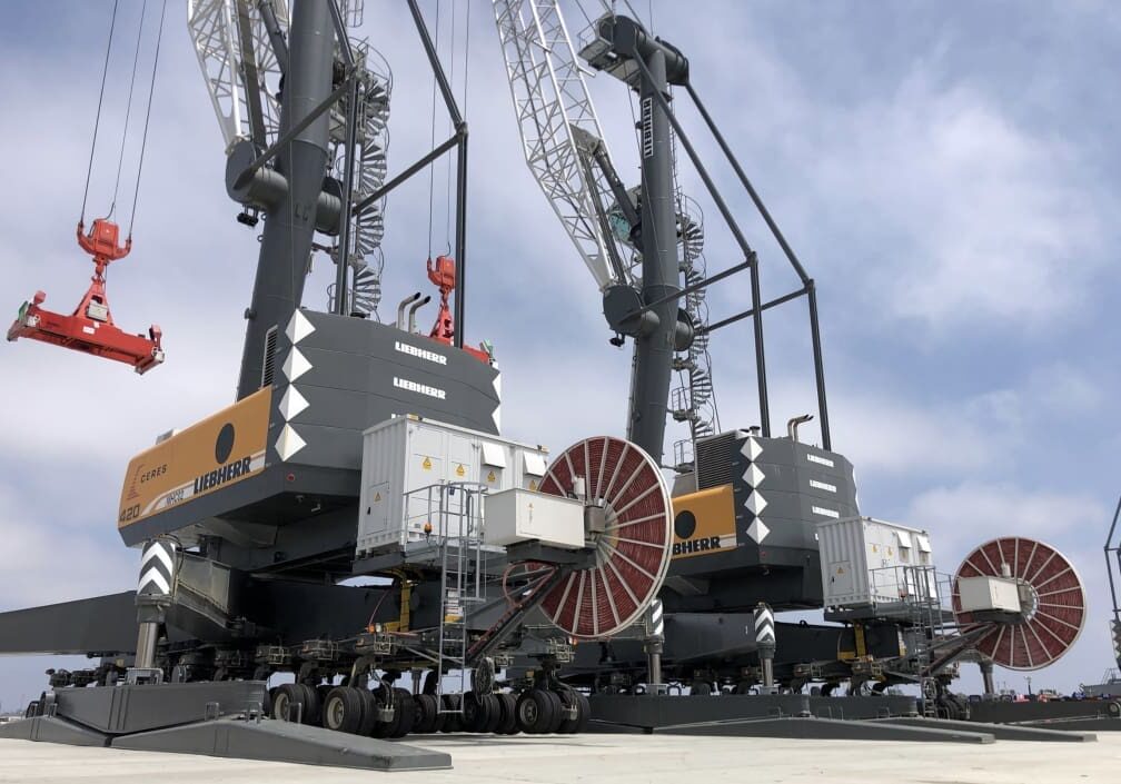 Three large Liebherr port cranes with extended booms and red hooks stand on a concrete surface under a partly cloudy sky, evoking the bustling activity of dock talks often held at year-end 2021.