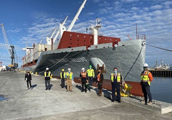 Eight people in safety gear stand in front of a large docked cargo ship on a sunny day at the port, engaging in some Dock Talk about the year's accomplishments before bidding farewell to 2021.