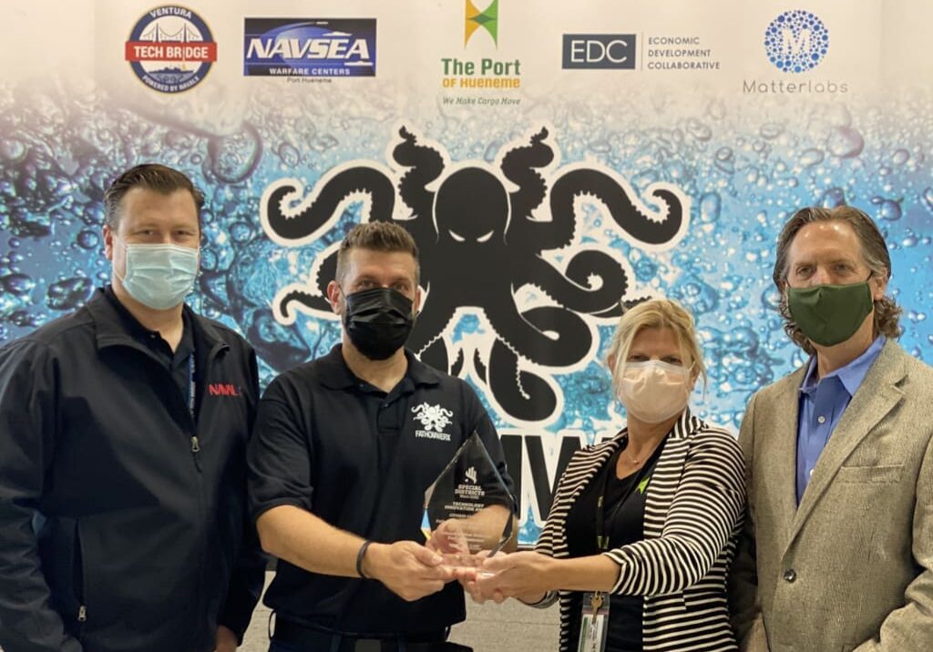 Four individuals are standing in front of a banner with logos, holding and presenting an award. All are wearing masks and business attire at the 2021 Year-End Dock Talk event.