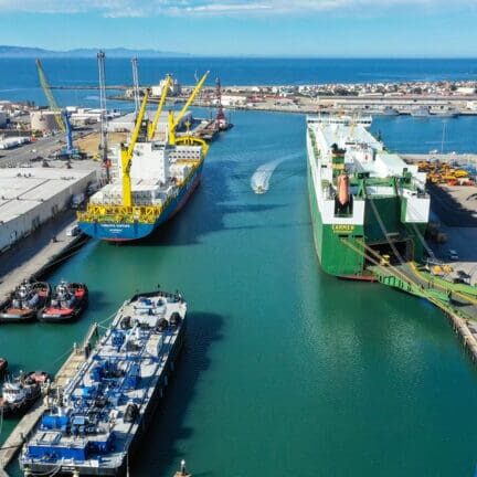 Aerial view of a busy shipyard with two large cargo ships docked along a narrow waterway, surrounded by industrial facilities and equipment. The clear blue sky and distant land on the horizon give an almost homely feel to this bustling maritime hub.