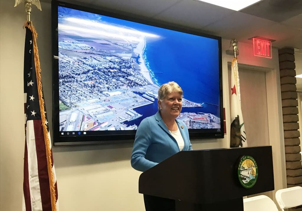 A woman stands at a podium with a large aerial view image of a coastal city on a screen behind her, reflecting the key points of Dock Talk 2021. An American flag and a California state flag are positioned on either side of the podium, setting the stage for the Year End review.