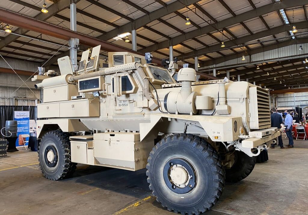 A large beige armored military vehicle is displayed in a warehouse setting with equipment and booths in the background, reminiscent of a Dock Talk feature from 2021.