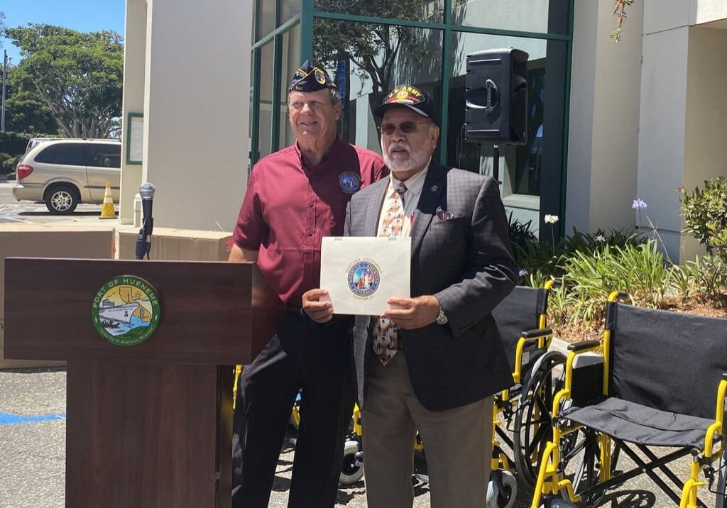 Two men stand next to each other outside a building. One is holding a certificate and the other is standing behind a podium with a seal that reads "City of Pico Rivera." Several wheelchairs are visible, marking the significant achievements discussed during the 2021 Year End Dock Talk event.