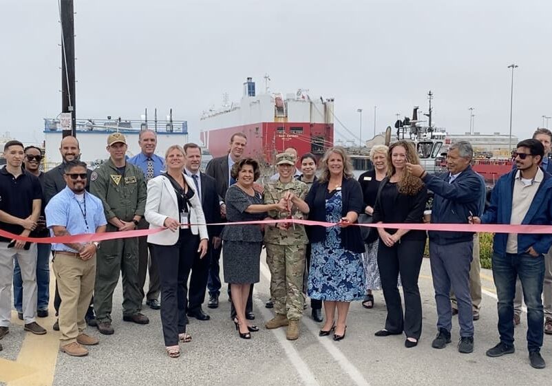 At the Year End 2021 ribbon-cutting ceremony in the port area, a group of people, including officials and military personnel, stand together with a large ship and cranes in the background. The event marked an important milestone discussed extensively in recent Dock Talk editions.